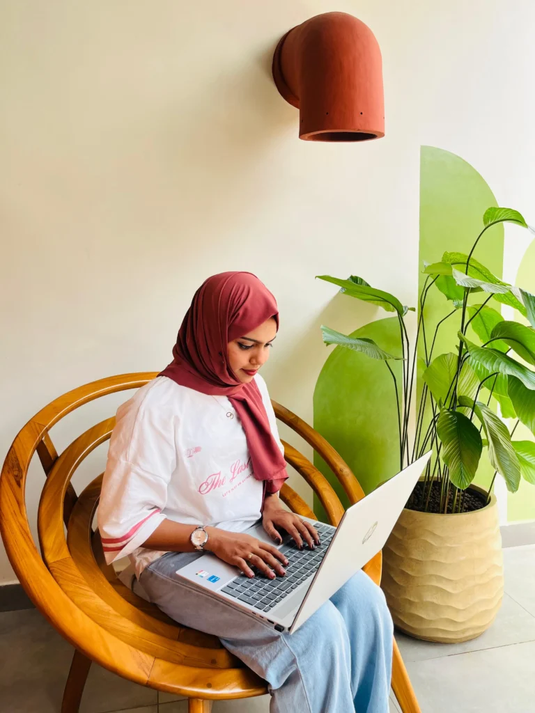A woman in a hijab sitting on a wooden chair and working on a laptop indoors.